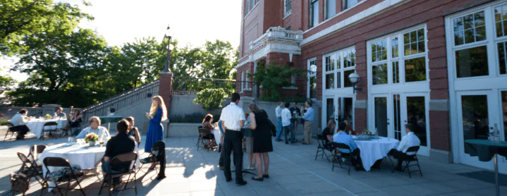 Several people socializing, standing and sitting at tables outside of a building.